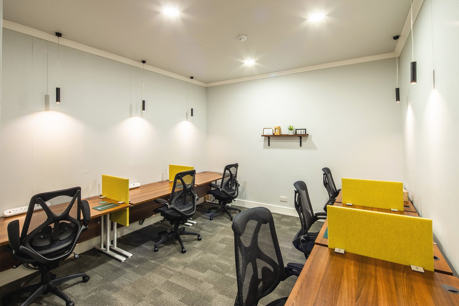 This modern office space in Scott House features six workstations with black mesh chairs and wooden desks. Each desk is paired with a yellow divider. The room boasts ample lighting from ceiling fixtures, and a small decorative shelf adorns the white wall.