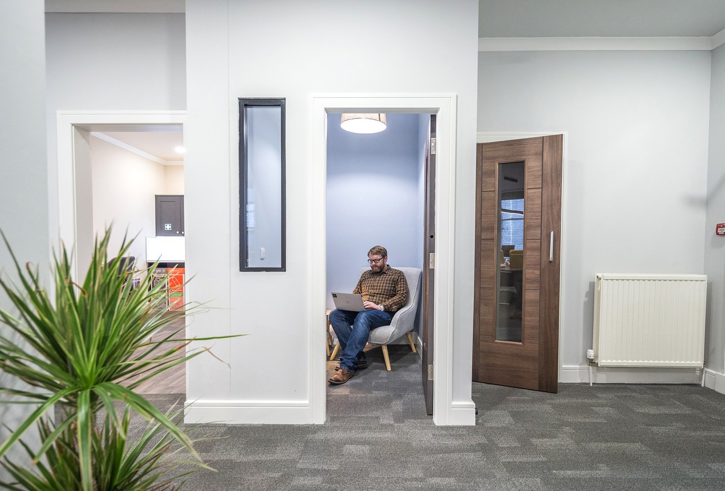 A person sits on a chair in a small, open-walled room on George Street, using a laptop. The space has light gray walls and a plant in the foreground. Two doors are visible, one open and one closed, enhancing the offices contemporary charm.