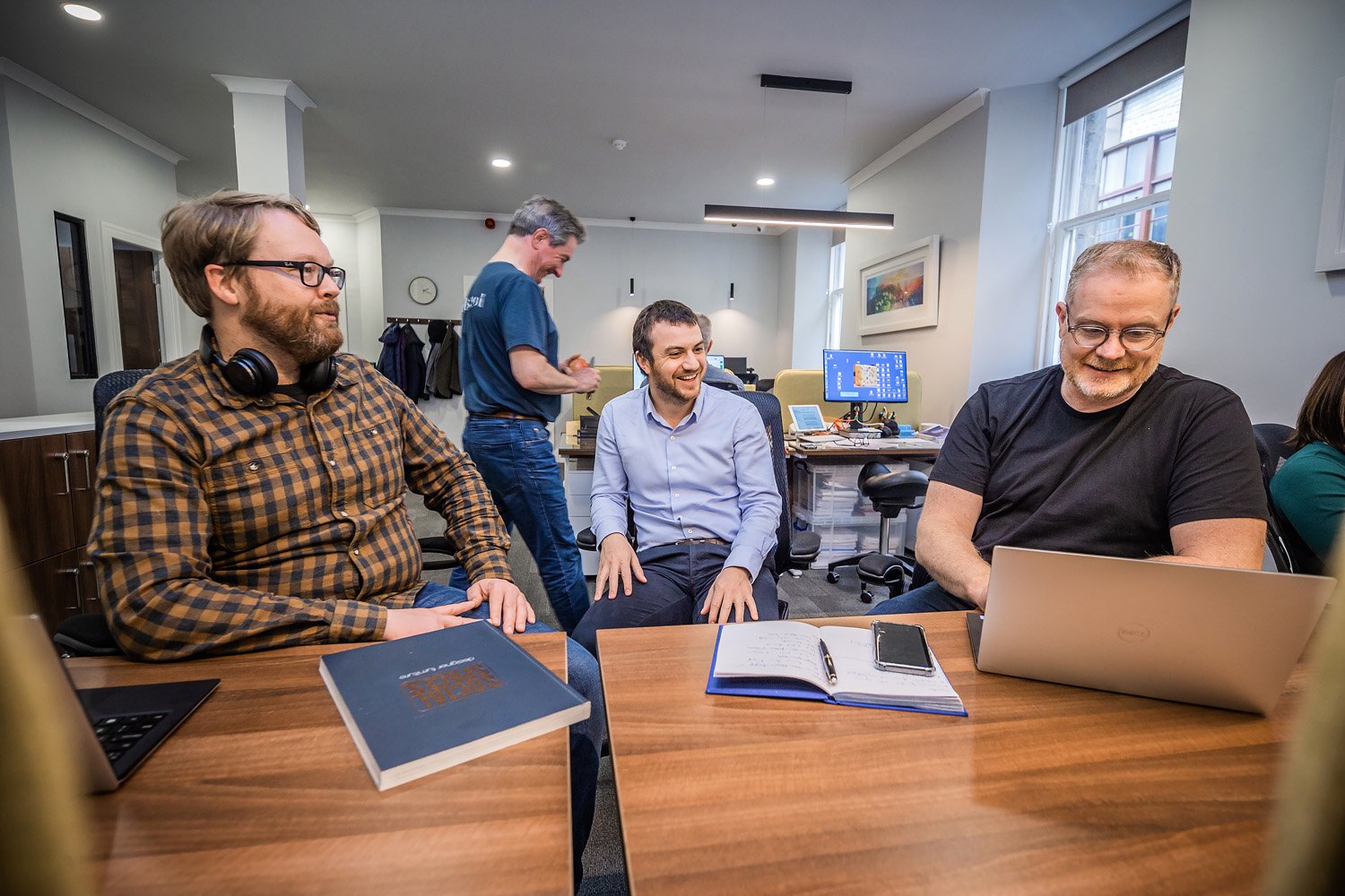 In a sleek York Place office, four men are engaged in a meeting. One smiles with his laptop, another listens with a notebook, the third adjusts a chair while the fourth wears headphones. A wall clock and desktop computer complete this modern scene.