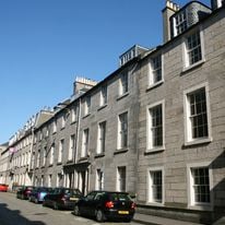 A row of gray stone buildings with tall windows lines Hill Street under a clear blue sky. Several cars are parked along the street in front of the buildings, enhancing the quaint charm of this bustling neighborhood.
