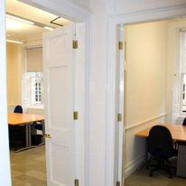 Located on Hill Street, this office space features two open doors leading to separate rooms. Each room contains wooden desks with black chairs and windows letting in natural light. The walls are painted white, and the floors are covered in neutral-toned carpet.
