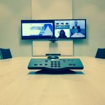 The Hill Street conference room features a long table with a remote control centered on it. Two wall-mounted screens display a video conference with several participants. The room exudes modern minimalism and is perfect for productive meetings.
