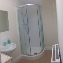 A small bathroom on Hill Street features a corner shower enclosure with glass doors. A wall-mounted sink with a mirror sits to the left, and a radiator is on the right. This compact space boasts beige walls and flooring, combining functionality with style.