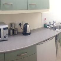A kitchen counter on Hill Street features a silver microwave, a stainless steel electric kettle, and three metal canisters. To the right is a sink with a tap, surrounded by light green cabinets both above and below.