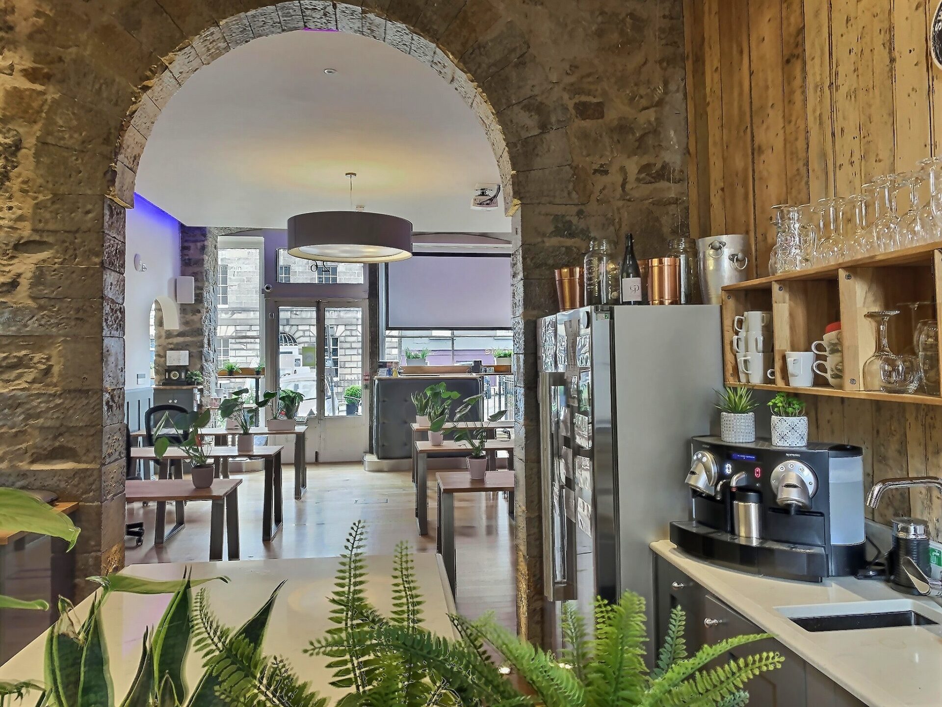 Nestled at 2 Walker Street, this cozy café boasts a charming interior with stone walls and wooden accents. A coffee machine on the right is surrounded by potted plants, while an archway leads to a seating area where tables and chairs bask in natural light from large windows.