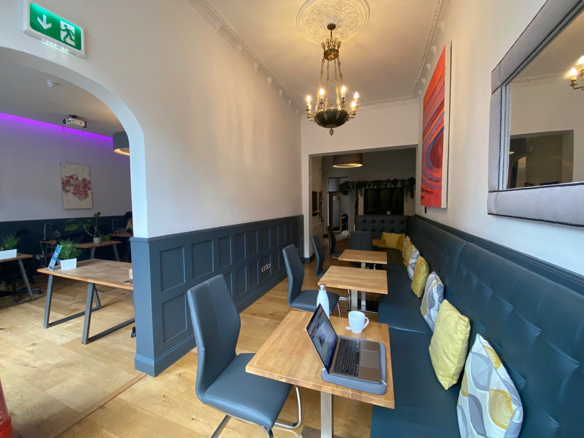 Located at 2 Walker Street, this contemporary cafe interior boasts a blend of blue and wooden decor. Tables feature chairs with colorful pillows, while a laptop and cup rest on one. A chandelier and modern art add elegance, an exit sign offers direction, and wood flooring completes the look.