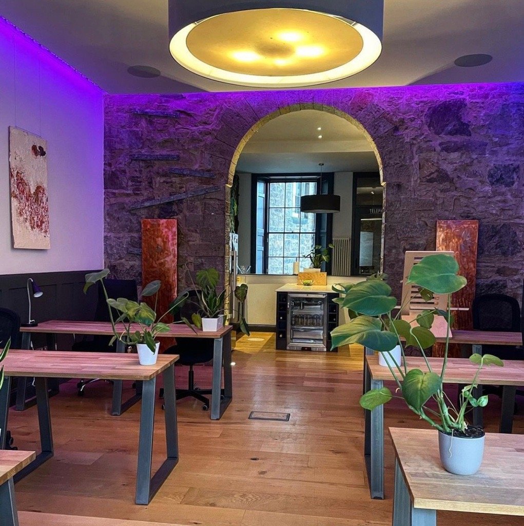 Located at 2 Walker Street, this modern office space features wooden desks, potted plants, and stone accent walls. A curved archway leads to a kitchenette, with purple LED lighting highlighting the stone walls and a large circular ceiling light adding warmth to the room.