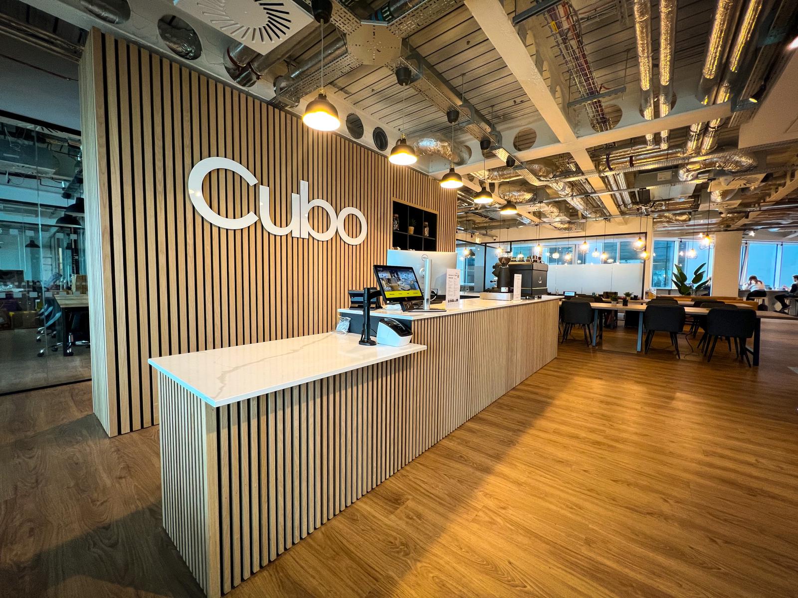 A modern office interior in Edinburgh features a reception desk with a Cubo sign. The space boasts wooden flooring, striped wooden panel walls, and exposed ceiling pipes. In the background, tables and chairs create an inviting workspace atmosphere.