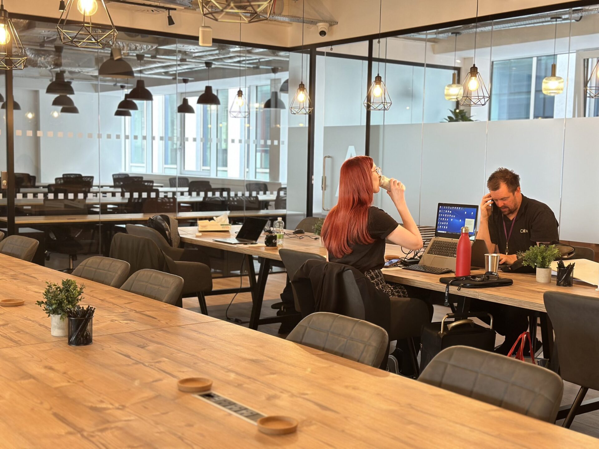 A woman with red hair sits at a large wooden table in a modern Edinburgh office, using a laptop and sipping from her water bottle. A man works on a laptop across from her, surrounded by hanging lights and large windows that offer stunning city views.