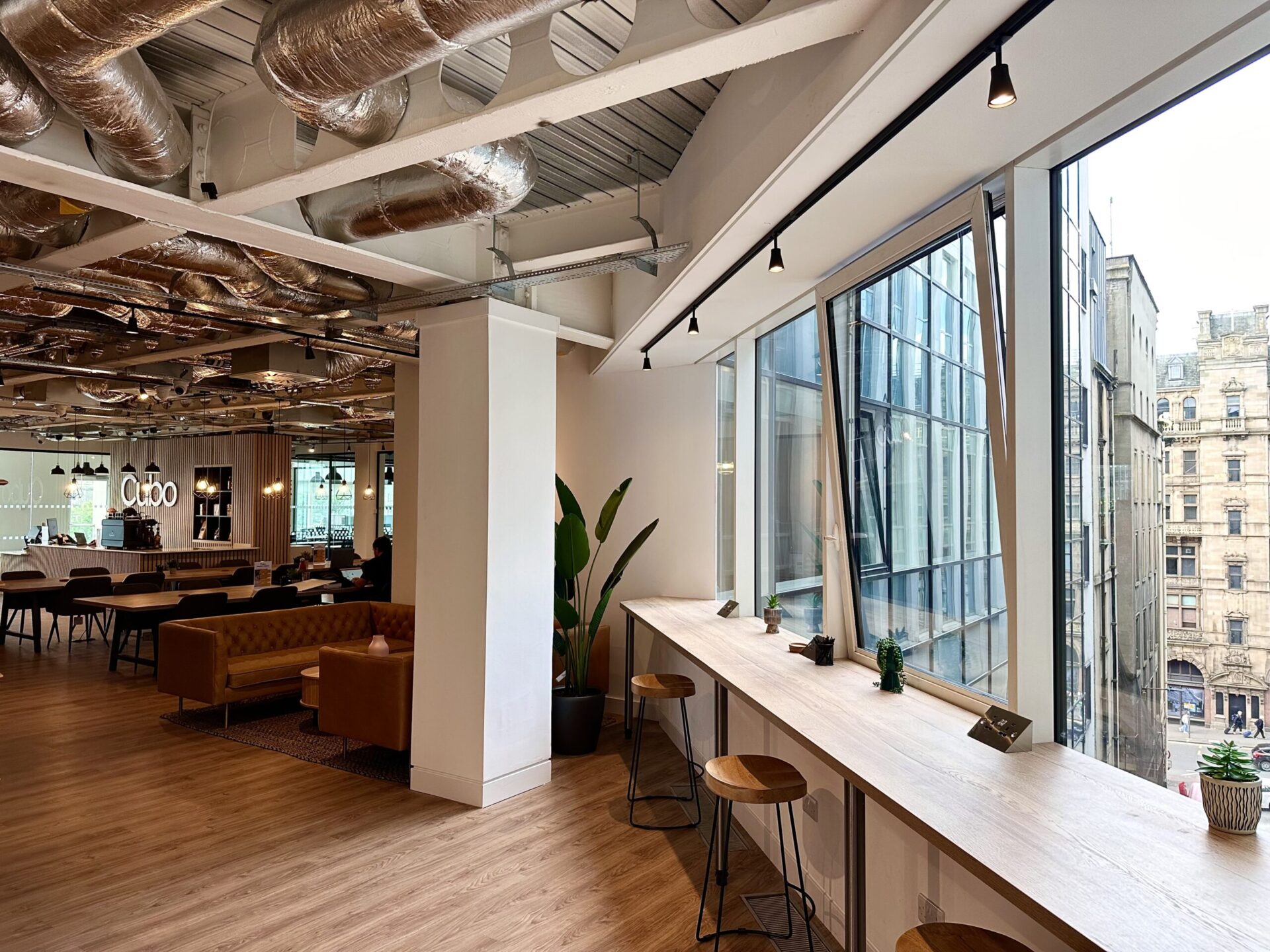 Modern office space in Edinburgh with large windows and city views. Features a long wooden counter with stools, plants, ceiling pipes, and a seating area with brown sofas in the background. Bright, airy atmosphere filled with natural light.
