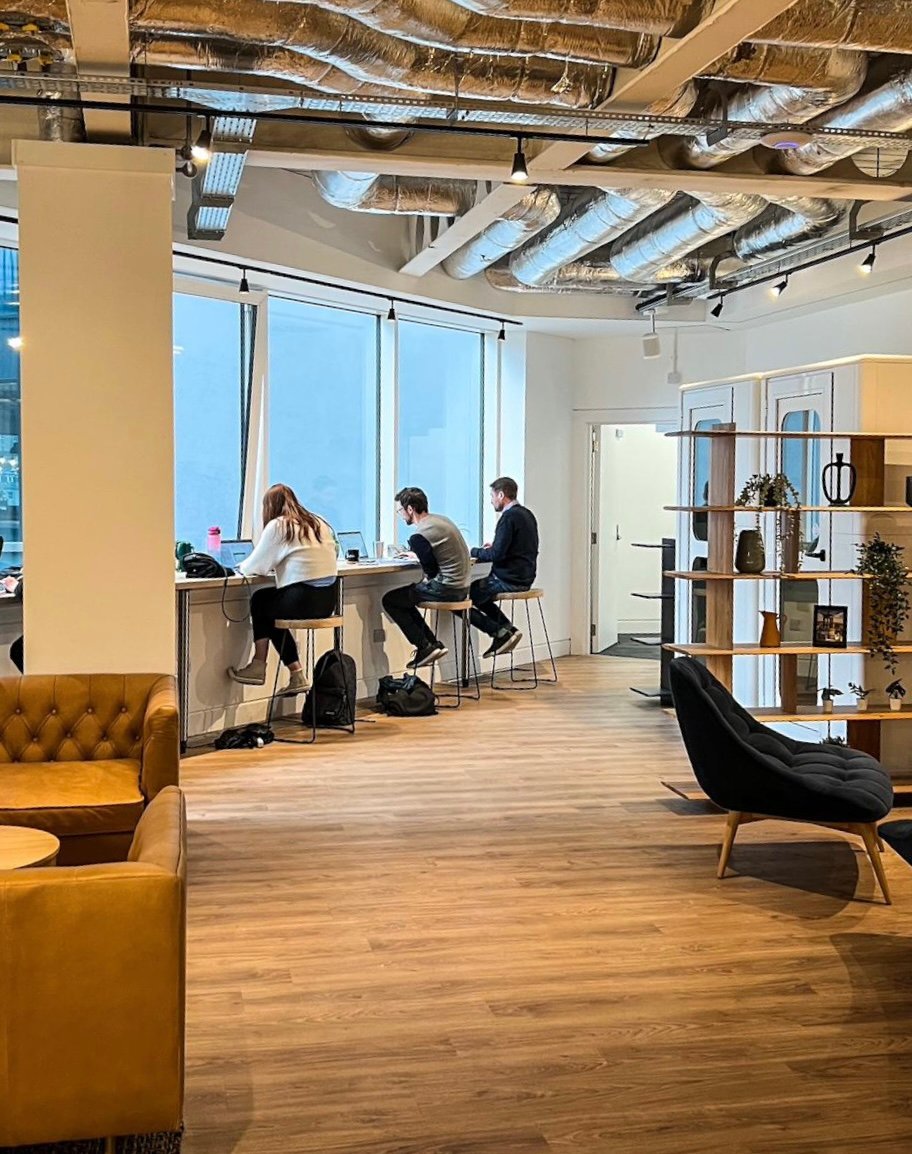 A modern office space in Edinburgh features three people working on laptops at a counter by large windows. The interior boasts exposed ceilings, a wooden floor, a yellow couch, a dark lounge chair, and a bookshelf adorned with plants and decor.