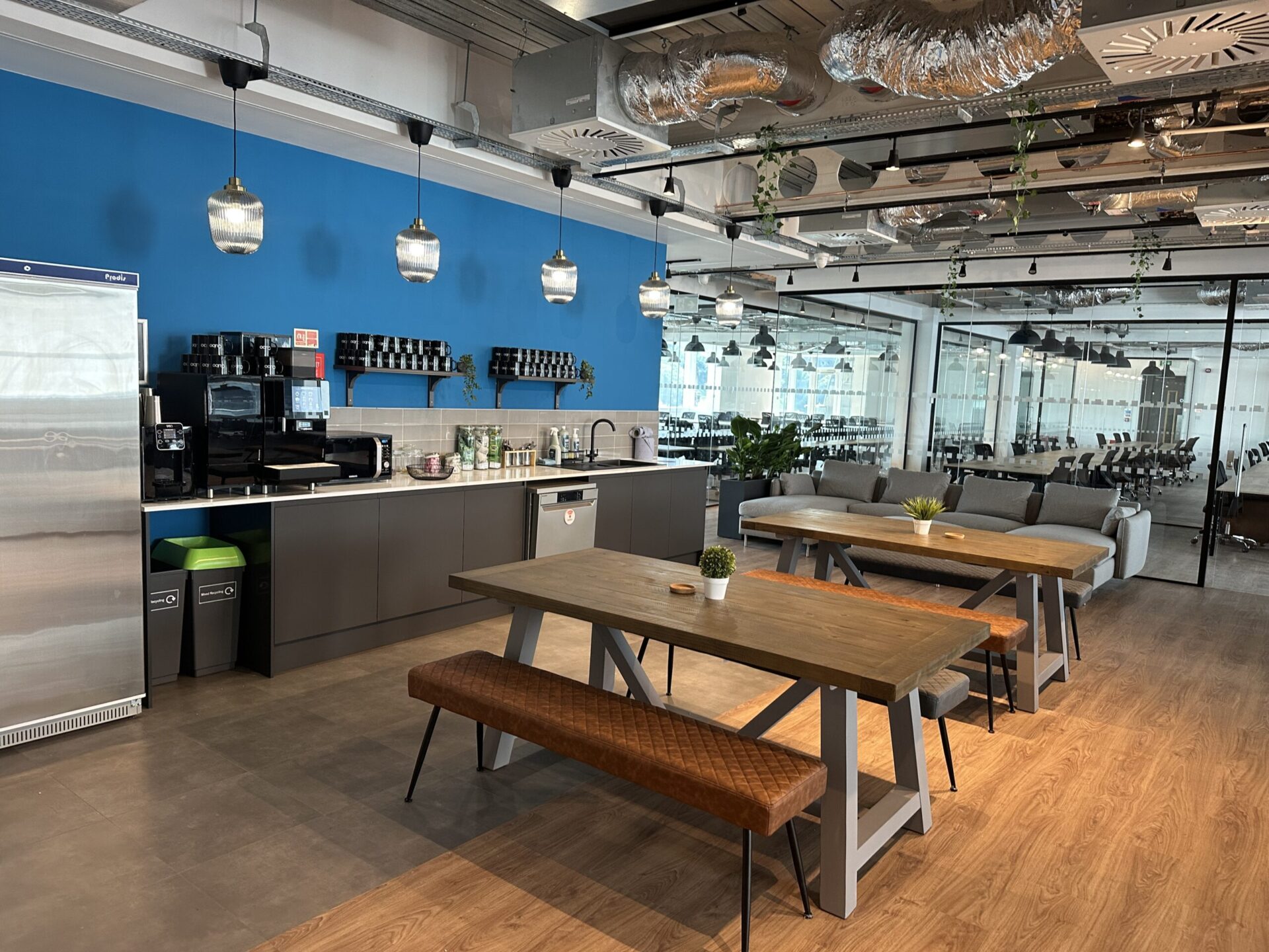 Modern office kitchen area in Edinburgh featuring wooden tables and benches, a blue accent wall, stainless steel appliances, and pendant lights. The space includes plants, a water cooler, and offers a view of a glass-walled conference room in the background.