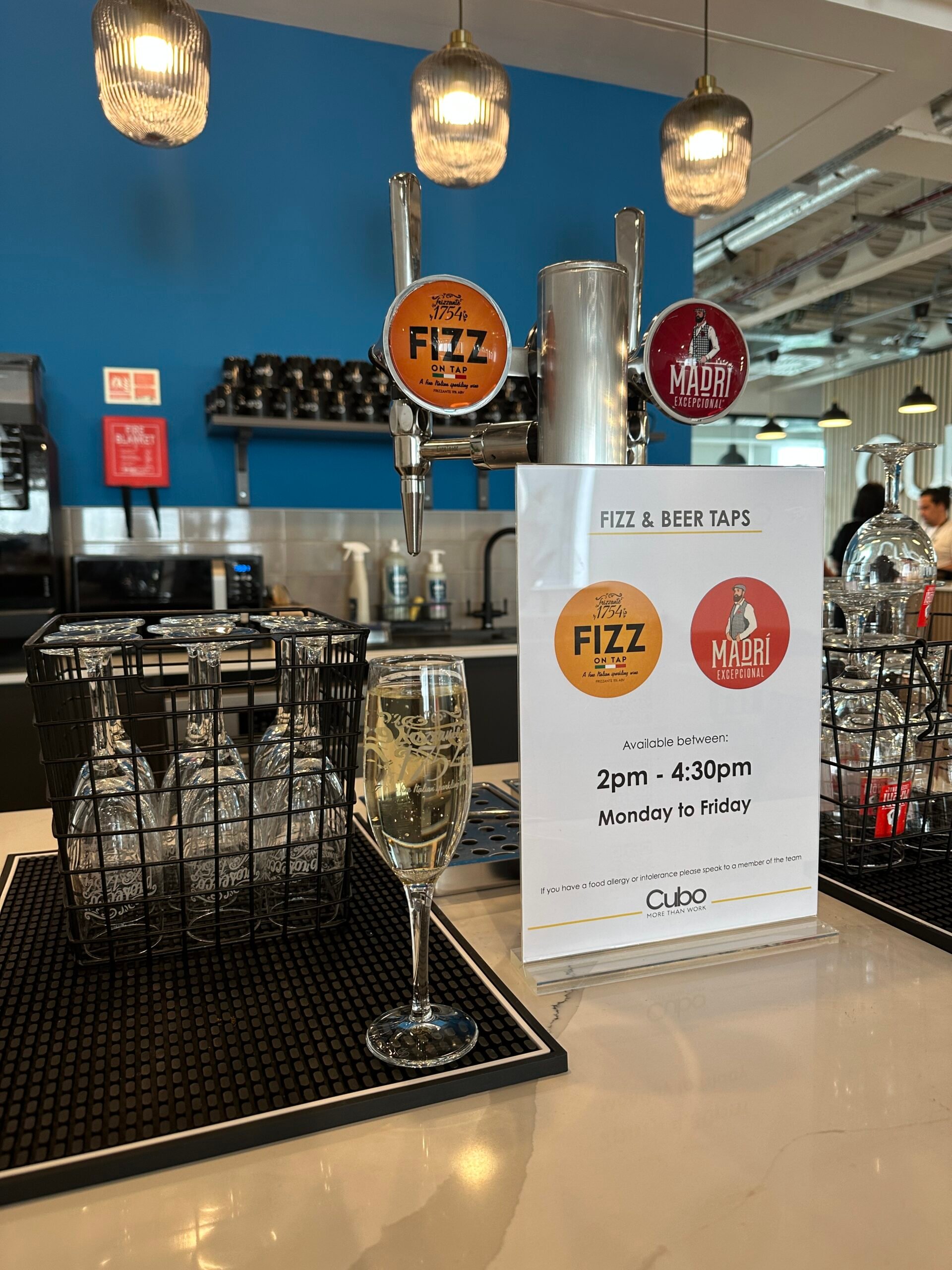In a stylish Edinburgh bar area, beer taps labeled FIZZ and MADRI stand ready. A glass of sparkling wine gleams on the counter. Behind it, a modern kitchen hums. A sign notes beer is available from 2pm to 4:30pm, Monday to Friday.