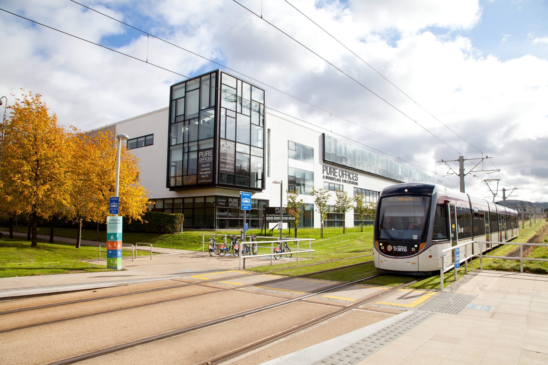 A modern tram glides through Edinburgh Park, its tracks hugging a building labeled PURE OFFICES. Autumn trees line the path, and bicycles rest near a rack. The partly cloudy sky enhances the bright and open atmosphere.