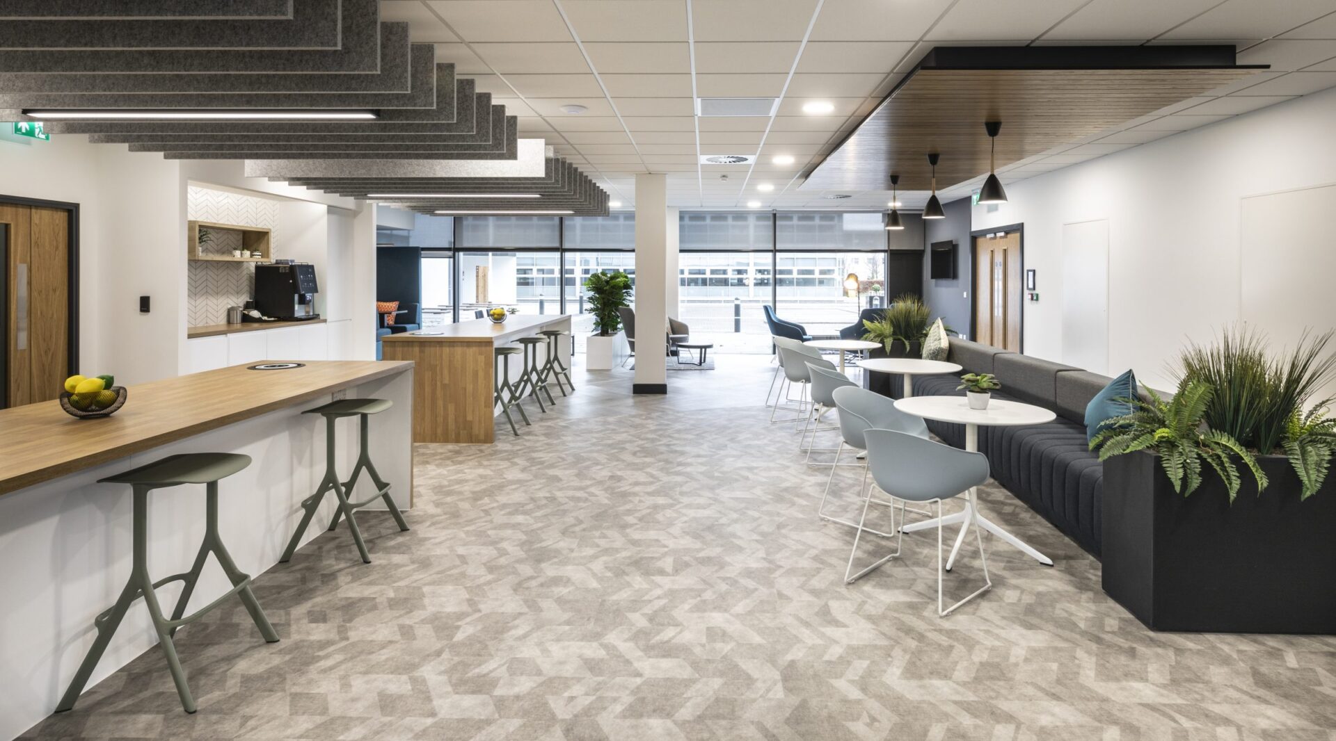 In Edinburgh Park, the modern office lounge boasts bar stools at a high counter, small tables with chairs, and a cozy sofa. Plants add an inviting touch, while large windows flood the space with natural light. Gray acoustic panels line the ceiling over a geometric-patterned floor.