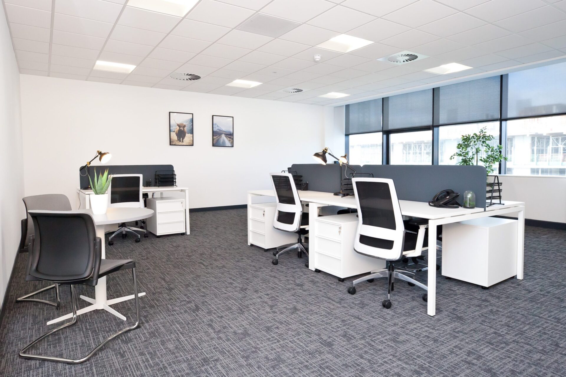 Located in the heart of Edinburgh Park, this modern office space features white desks, black chairs, and a round table with a plant. Two framed pictures adorn the white wall. Large windows with blinds invite natural light, while dark gray carpeting adds a touch of elegance.