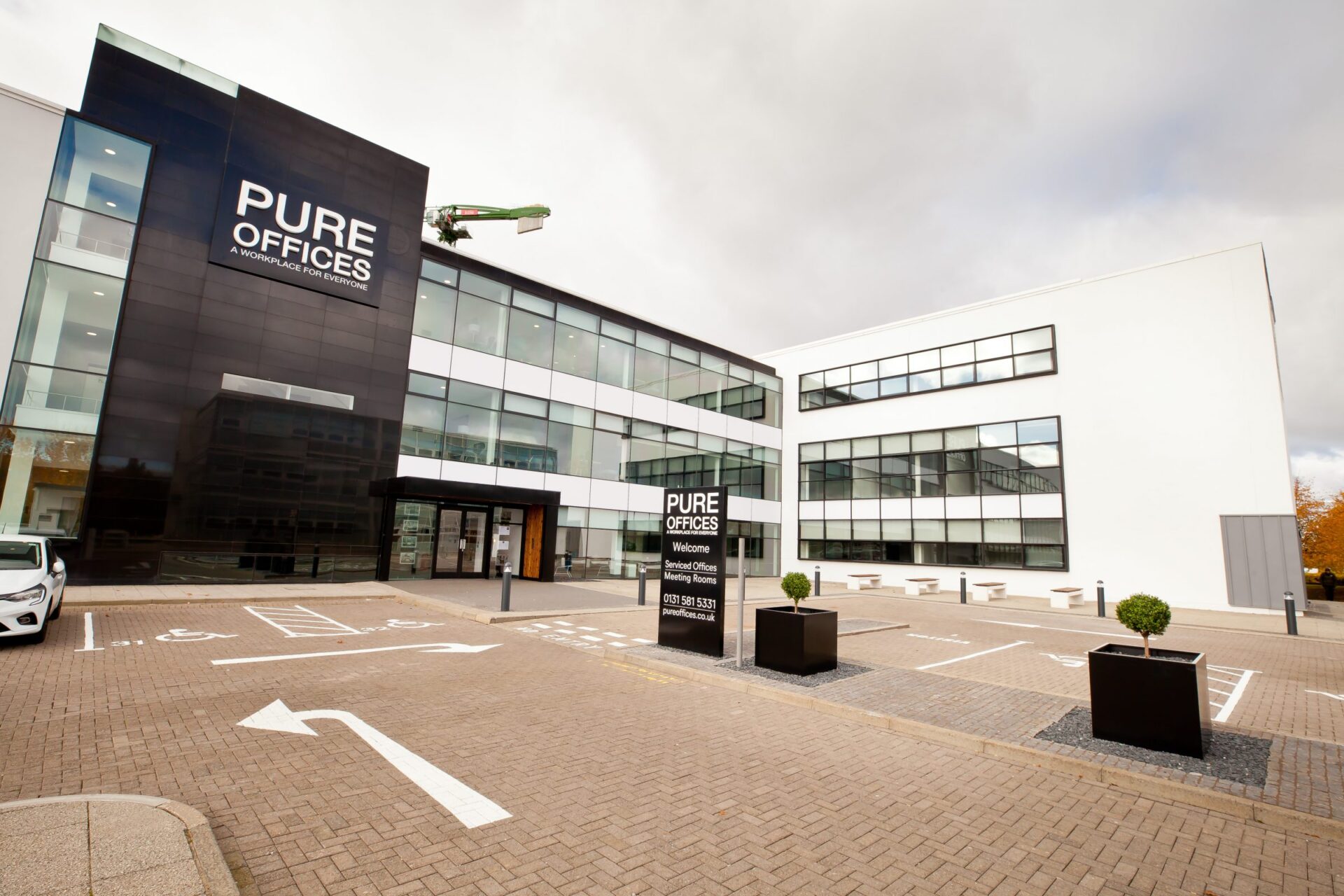 Modern office building with a sleek black and white exterior located in Edinburgh Park. The signage reads Pure Offices with contact details. The area features parking spaces, outdoor seating, and large windows. A worker on a lift is performing maintenance on the building.
