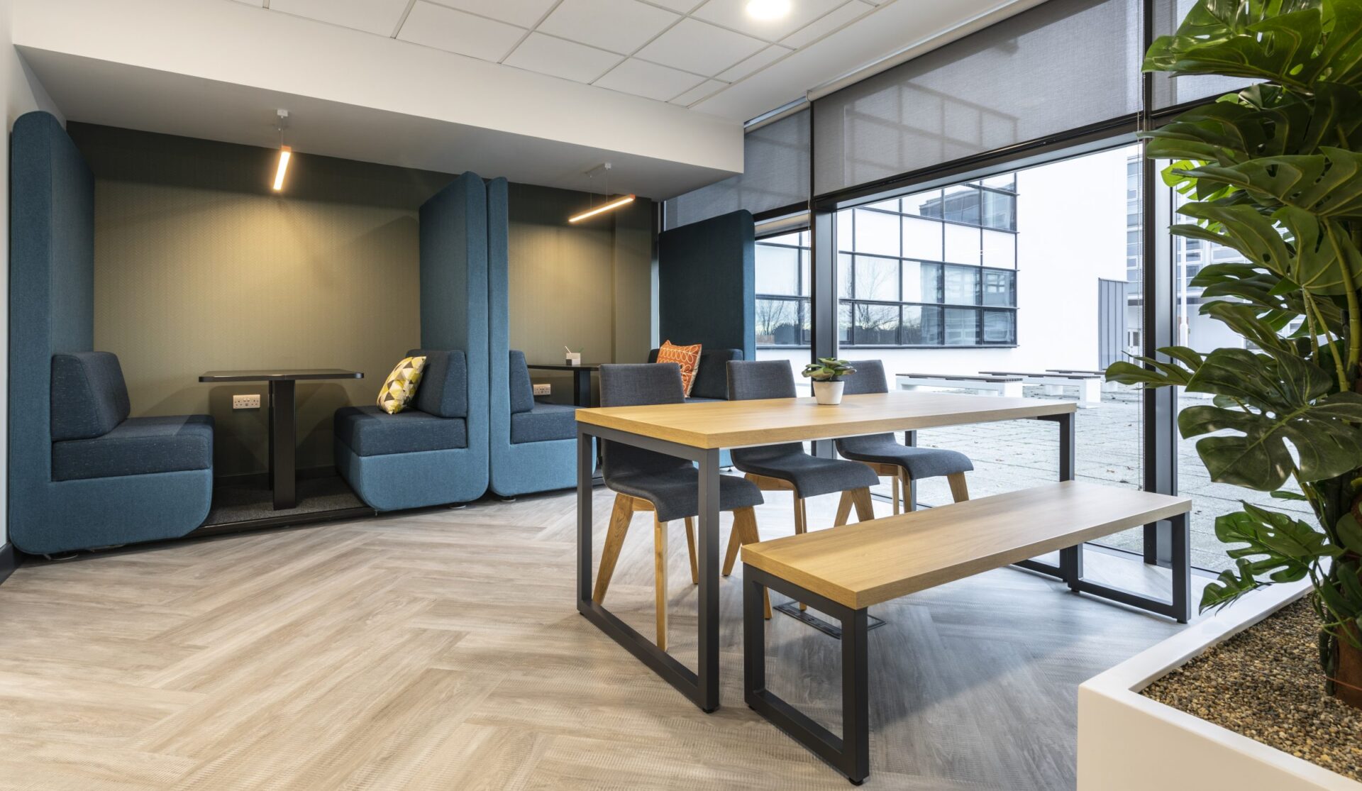 A modern office break area at Edinburgh Park features blue booth seating with cushions, a wooden table and benches, large windows with shades, and a corner plant. The light wood patterned floor complements the neutral walls, creating an inviting atmosphere for relaxation.