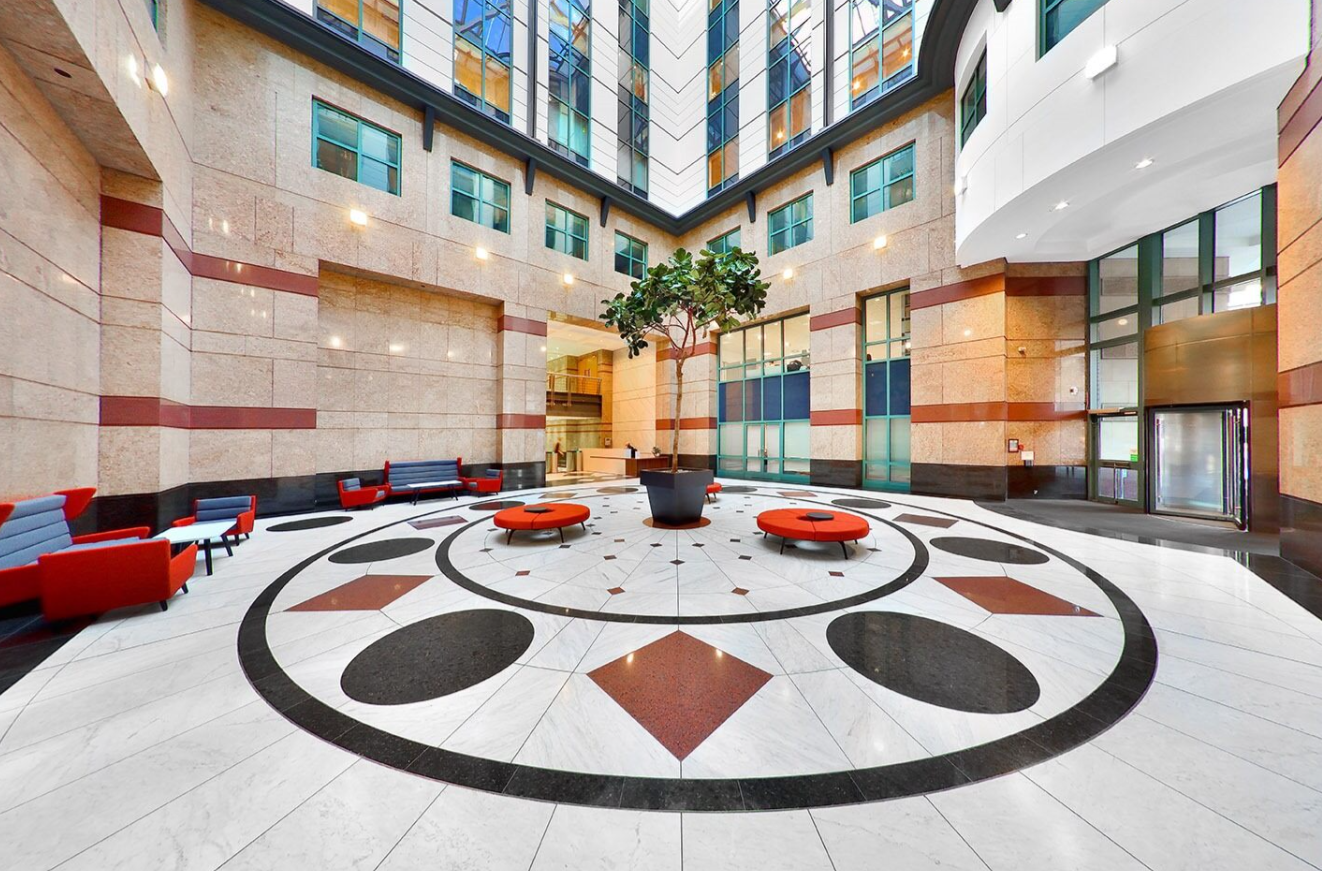 The Exchange Plaza boasts a spacious atrium with high ceilings, a patterned marble floor, modern seating, and a central potted tree. Surrounded by large windows and beige walls, its brightly lit with both natural and artificial light.