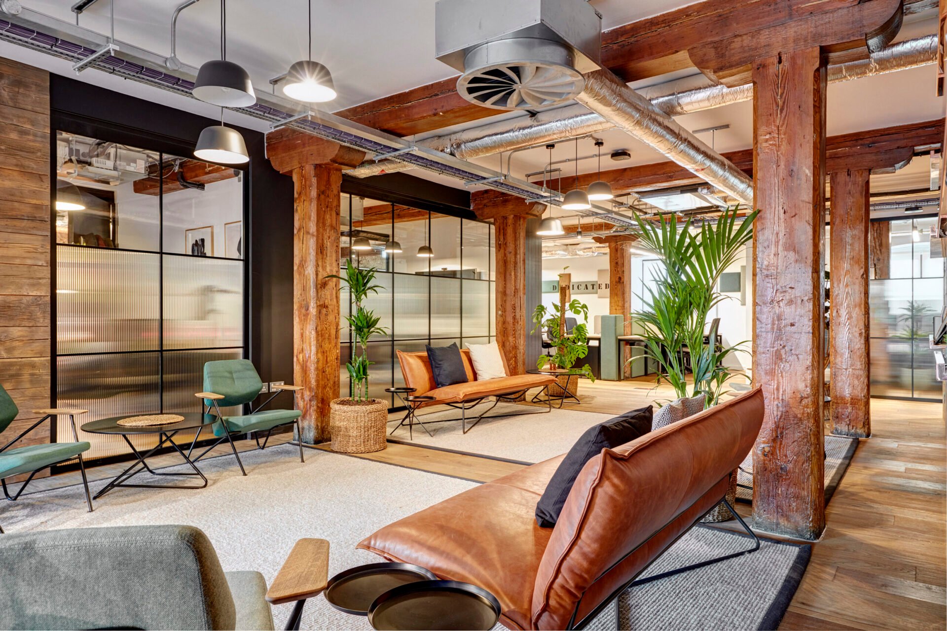 A modern office lounge in Edinburgh boasts wooden beams and industrial elements. It features leather sofas, plants, and round tables. Pendant lights hang from the ceiling, creating a cozy atmosphere. Large windows offer ample natural light.