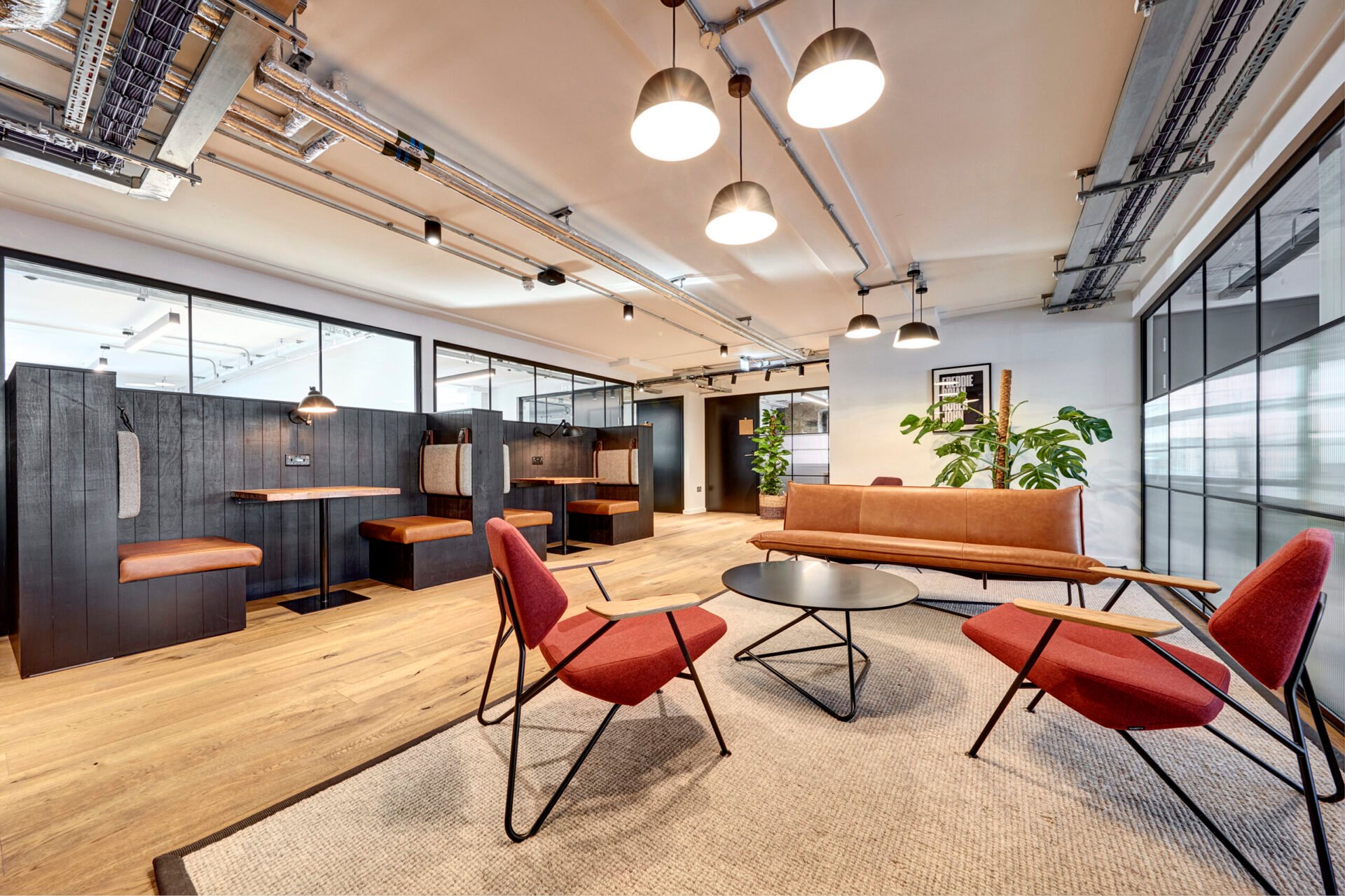 A modern office lounge in Edinburgh features red chairs, a round coffee table, and a tan sofa on a light rug. The black booth seating, wood flooring, and hanging ceiling lights add elegance, while large windows flood the space with natural light.