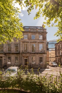 Historic three-story stone building with large windows, framed by leafy trees. A car is parked on the street in front. A modern glass building is partially visible in the background. Bright sunny day with a clear sky.