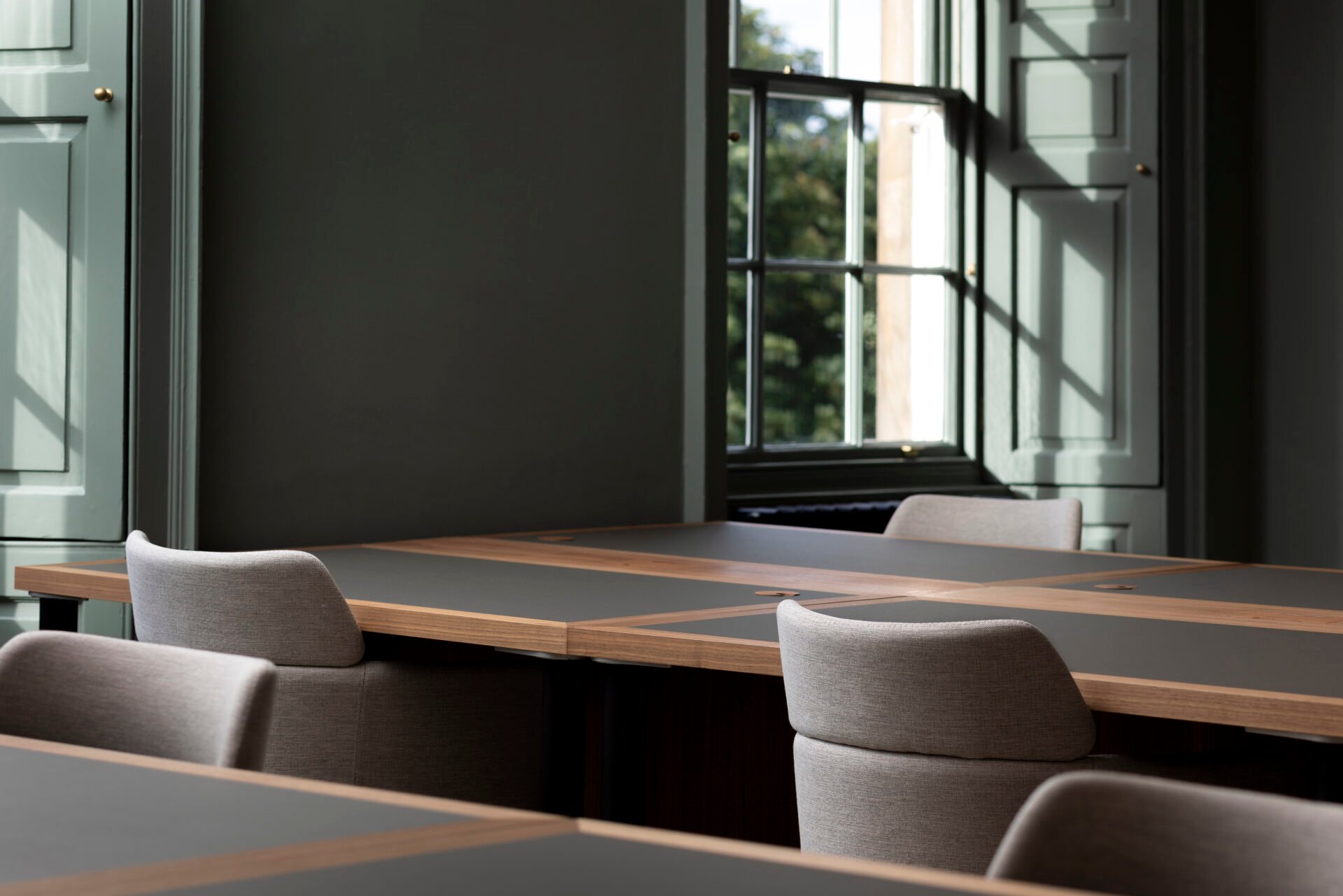 The modern conference room at 22 Queen Street features sleek wooden tables and light gray chairs. Sunlight streams through large windows, creating a tranquil atmosphere. The muted green walls complement the minimalistic and elegant design perfectly.