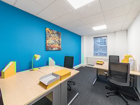 A bright office space on George Street features a blue accent wall adorned with a colorful abstract painting. Four desks come equipped with black chairs, yellow desk lamps, and folders. A large window floods the area with natural light.