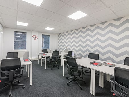 Located on George Street, this modern office space boasts several white desks and black chairs arranged in orderly rows. The walls showcase a stylish zigzag pattern in gray and white. With ample lighting and two windows gracing the left wall, it offers an inviting work environment.