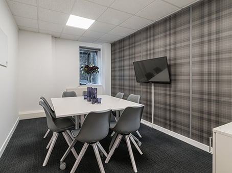 A modern conference room on George Street features a white rectangular table surrounded by six gray chairs. A flat-screen TV is mounted on a plaid-patterned wall. A small window with a bouquet of flowers adorns the ledge, and the floor is carpeted.