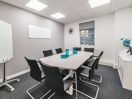 A modern conference room on George Street features a long table with eight black chairs, a blank whiteboard, and a window with a gray blind. The room is complete with a wall clock, a white cabinet holding blue flowers, overhead lighting, and dark carpeting.