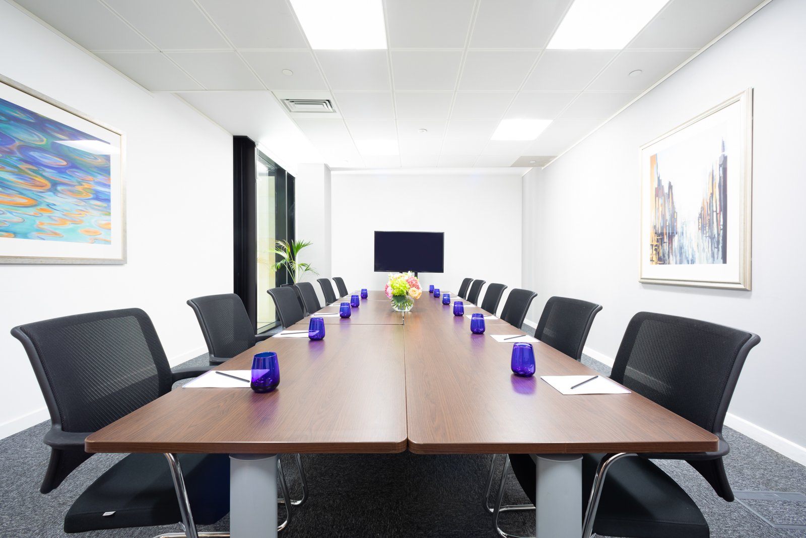 A modern conference room in Edinburgh City Centre features a long wooden table surrounded by black chairs. Blue glasses, yellow flowers, and notepads are neatly arranged. White walls adorned with abstract art and a flat-screen TV complete the sophisticated setting.
