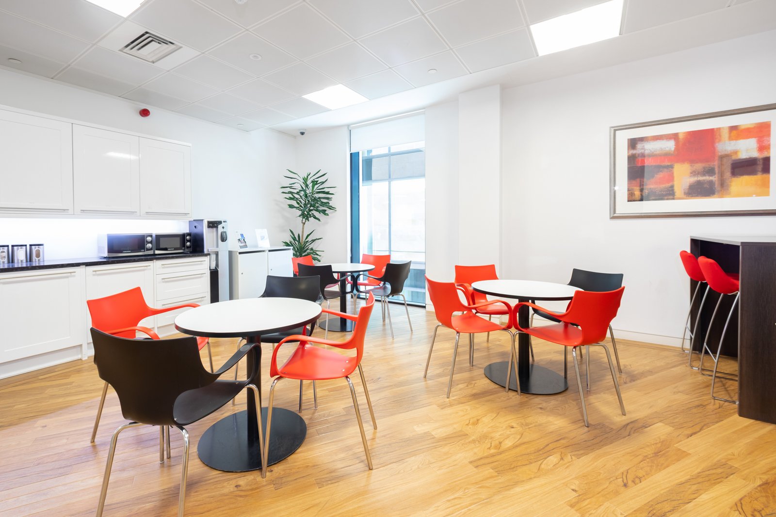 The modern office break room, reminiscent of Edinburgh City Centres chic vibe, features round tables with a mix of red and black chairs. To the left, a small kitchenette includes a microwave, coffee maker, and cabinets. A large abstract painting and potted plant complete the bright space.