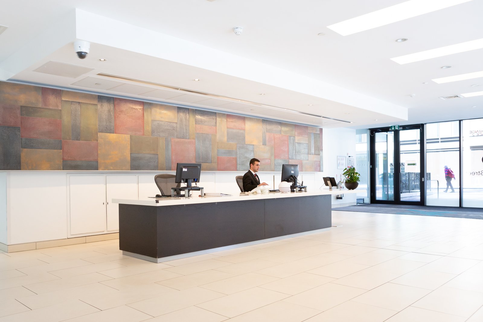 In a modern office lobby in Edinburgh City Centre, a person at the reception desk is busy working on two computers. The tiled floor complements the mural on the wall behind them, while large glass doors flood the space with natural light from outside.