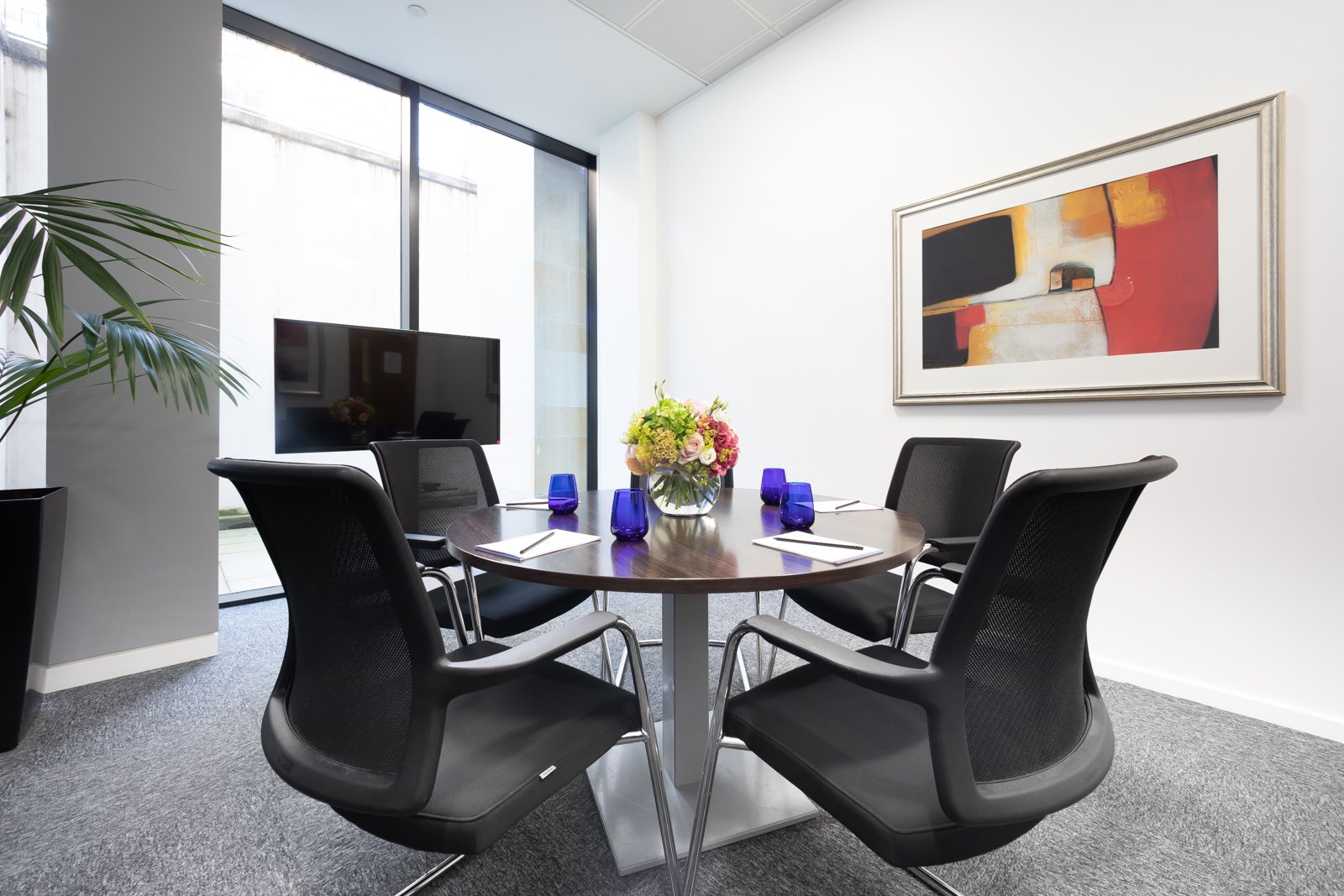 In the heart of Edinburgh City Centre, a modern meeting room features a round wooden table with four black mesh chairs. A colorful abstract painting graces the white wall, while natural light streams through a large window. A flat-screen TV and potted plant complete the spaces contemporary charm.