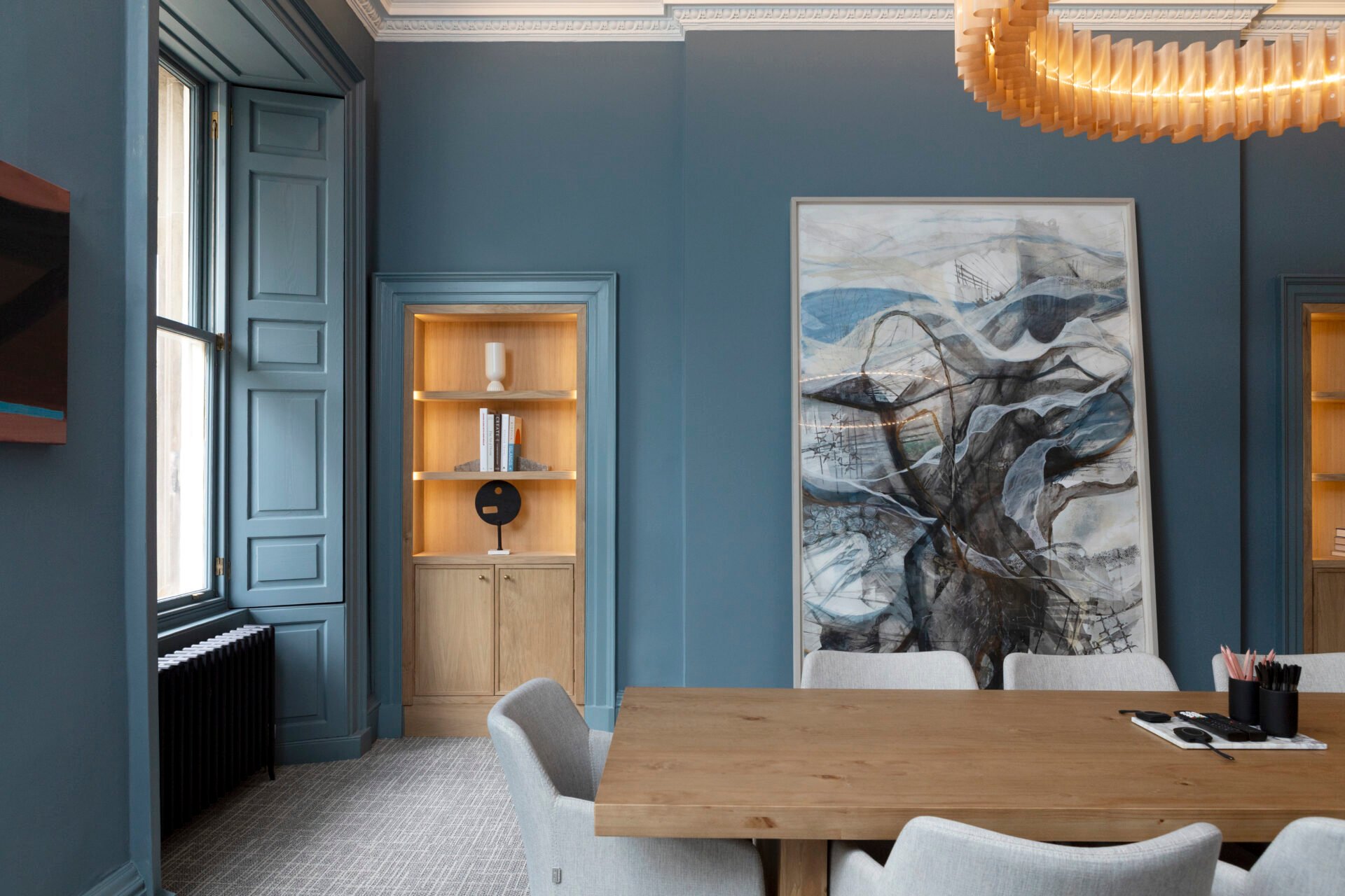 Located at 22 Queen Street, this modern meeting room features blue walls and a large abstract artwork. A wooden table is surrounded by gray chairs. Built-in shelves with decor and a large window add charm, while a contemporary chandelier hangs overhead, enhancing the stylish space.