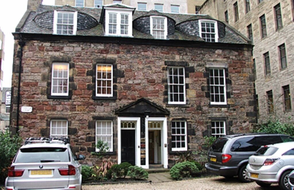 Thistle Court Business Centre is a three-story stone building with white-trimmed windows and a central entrance. The roof boasts dormer windows, while cars are neatly parked on the gravel driveway in front. Taller structures envelop this charming business hub.