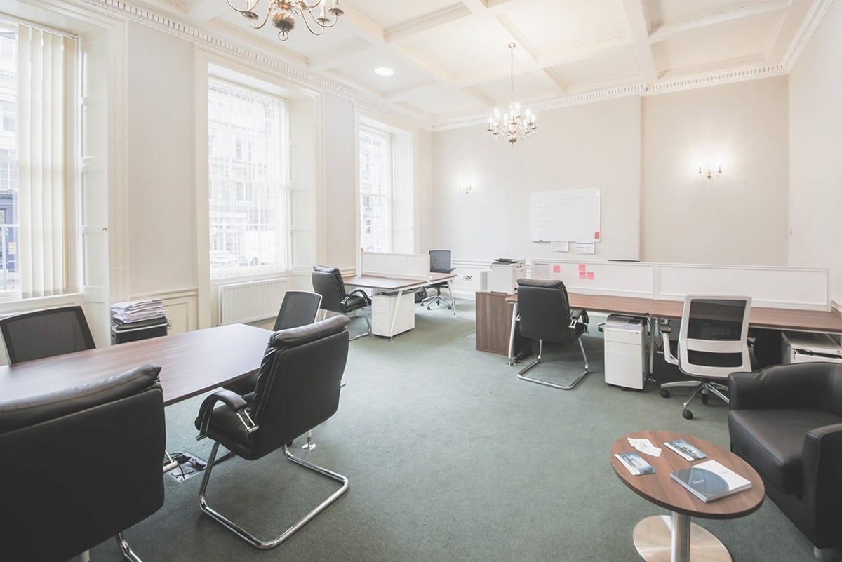 A bright office space on George Street features several black chairs and wooden desks. Large windows flood the room with natural light, illuminating a chandelier, a whiteboard on the wall, and a small round table with brochures.