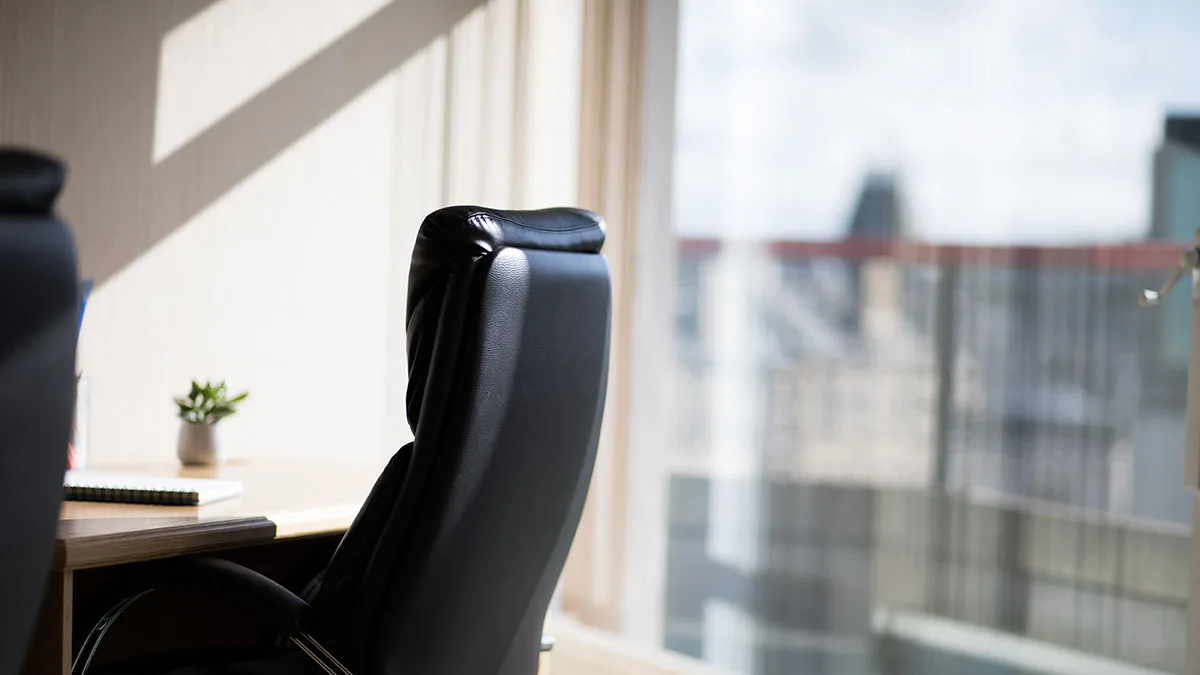 A sunlit office at York Place features a black leather chair facing a wooden desk. A small plant and an open notebook with a pen rest on the desk. Large windows reveal a blurred city skyline in the background.