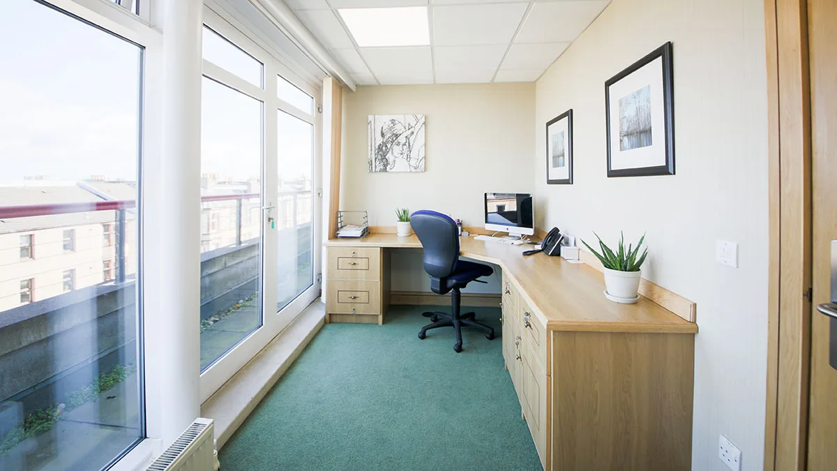 A small office space at Hopetoun Gate features a desk, chair, desktop computer, and telephone. Large windows flood the room with natural light. The green carpet and beige walls complement framed pictures and a potted plant that adds life to the desk.
