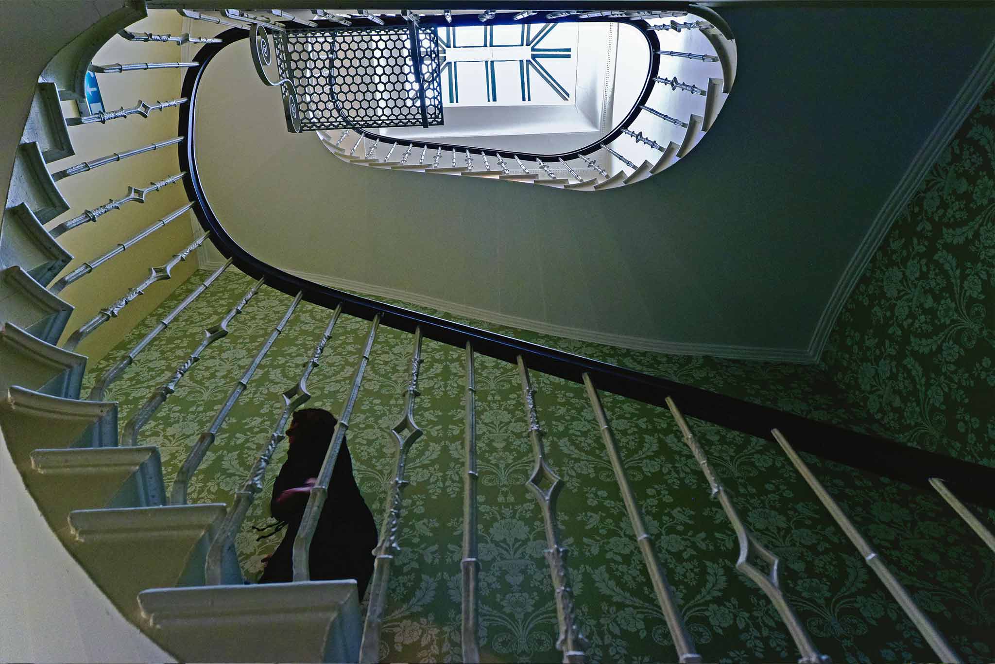 A spiral staircase with white railings and patterned green wallpaper graces the walls at 14 Albany Street. A person is ascending, partially obscured by the railing. The view is from below, looking up toward a bright skylight.