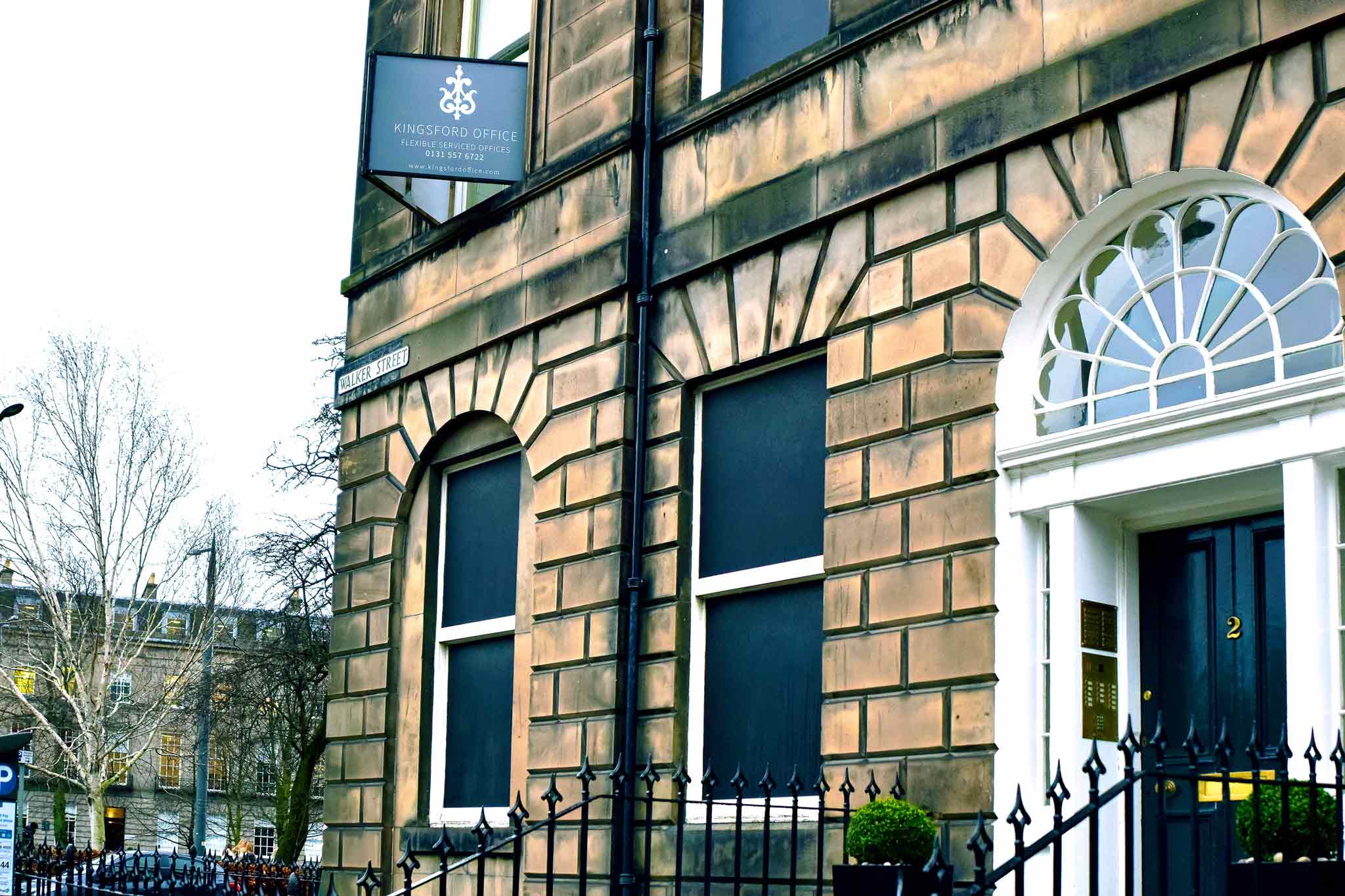 A stone building at 2 Walker Street proudly displays a sign for Kingsford Office above its arched door. It features black window coverings with white trim, while a black fence lines the foreground and bare trees grace the background.