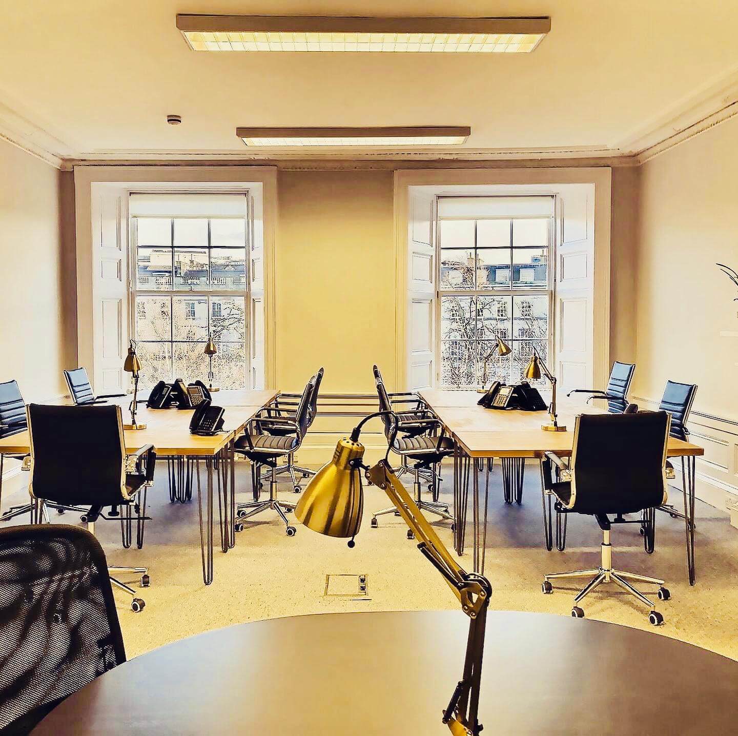 A brightly lit office at 2 Walker Street boasts several desks with black chairs arranged in rows. Each desk features a task lamp and other essentials. Two large windows at the back flood the space with natural light, offering a stunning cityscape view.