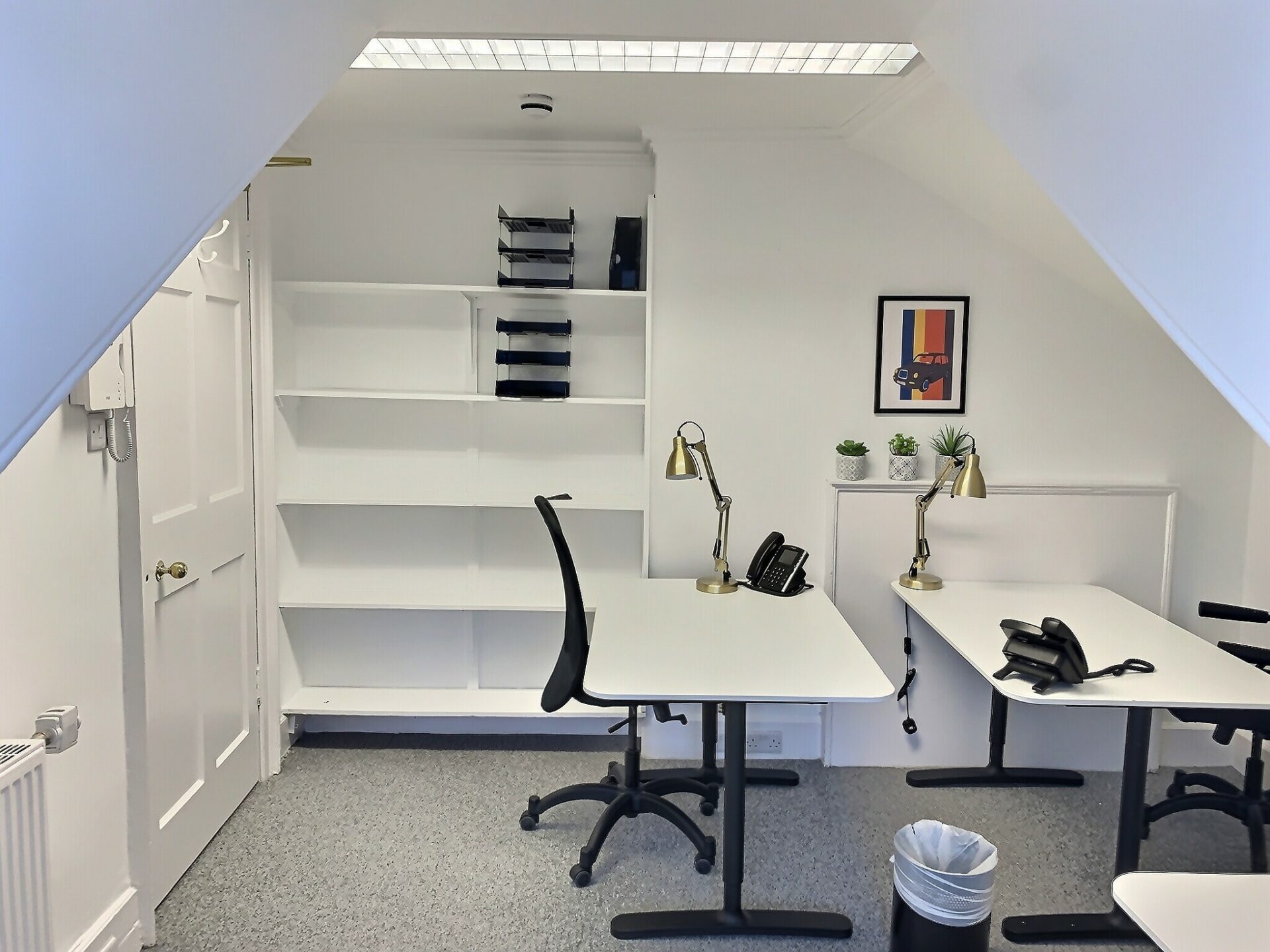 Located at 2 Walker Street, this small office features two white desks with black chairs. Shelves mounted on a white wall hold file organizers. A small framed picture and potted plants add charm, while a trash bin sits under one desk. Lighting is provided by a ceiling fixture.