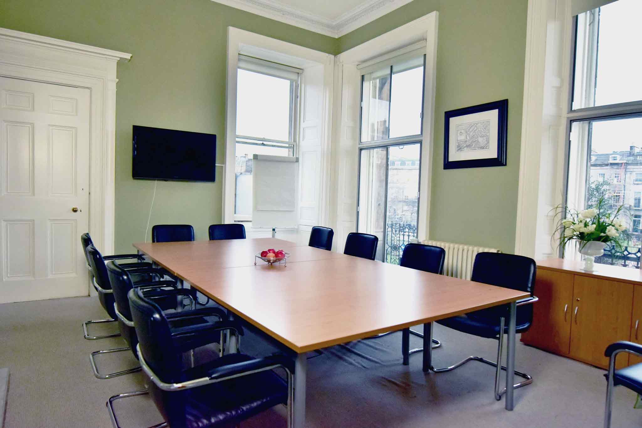 A conference room at 2 Walker Street features a large wooden table surrounded by black chairs. A TV is mounted on the green wall alongside a framed picture. Bright flowers adorn a cabinet near the spacious windows, and a small bowl of red apples sits invitingly on the table.
