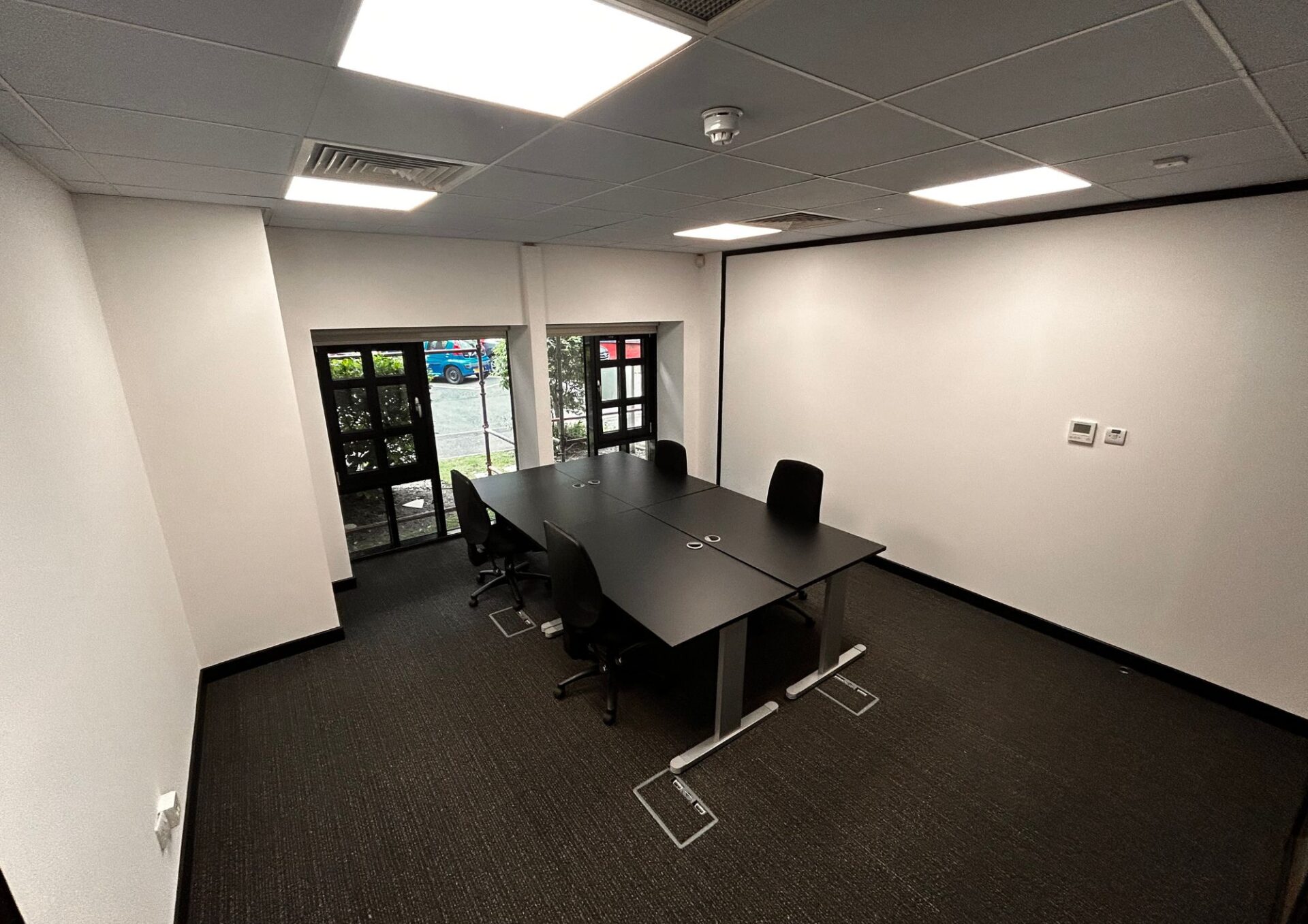 Office 1 Papermill Wynd features a modern workspace with a black table and six chairs, perfectly STACKed against large windows overlooking the parking lot. The room boasts white walls, a black carpet, recessed lighting, and convenient power outlets on the table.