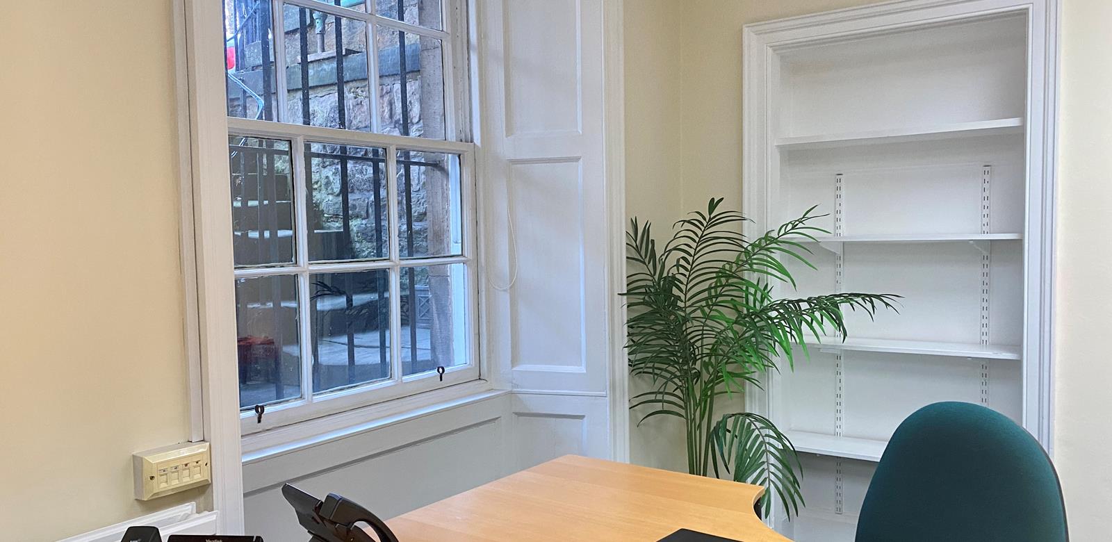 An office with a wooden desk and a green chair near a large window. A telephone and notebook sit on the desk. An empty white bookshelf and a potted plant are against the cream-colored walls.