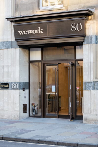 The entrance of a building at 80 George Street features a WeWork sign above sleek glass doors. The facade is made of stone, complemented by a tidy sidewalk. To the left of the doors, a smaller WeWork sign adorns the wall.
