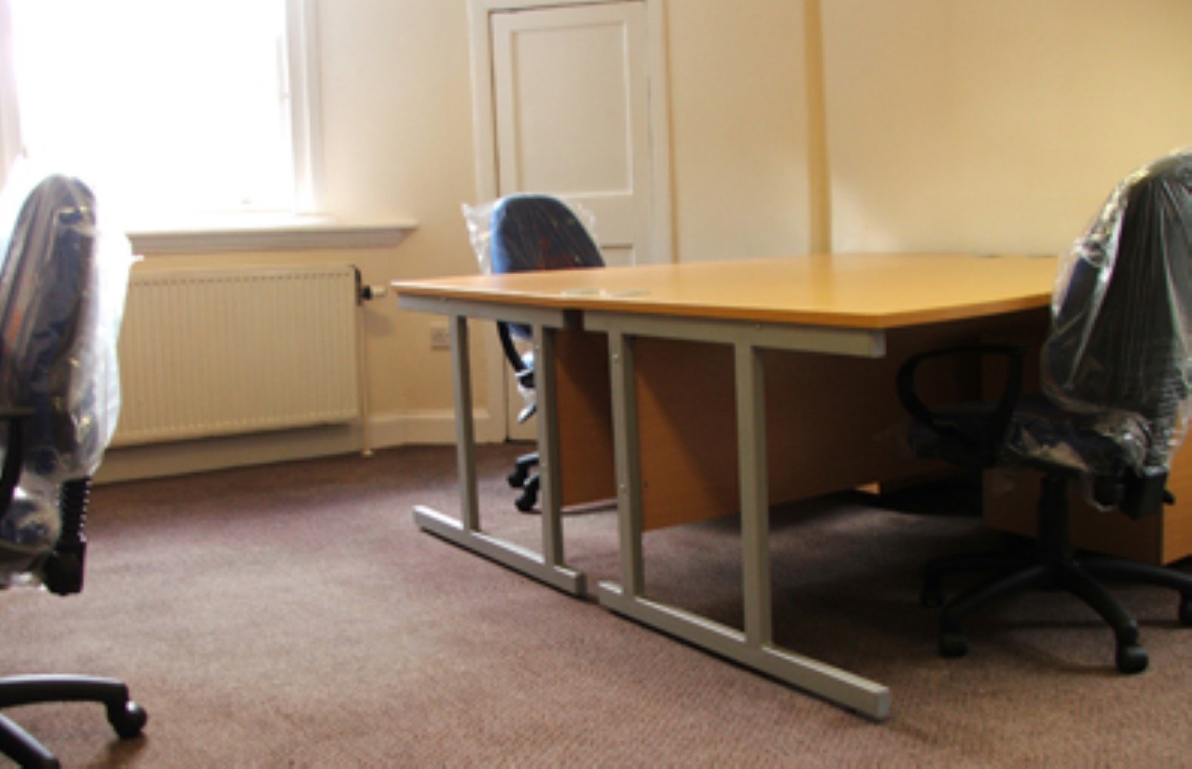An empty office space at Thistle Court Business Centre features a wooden desk and two blue office chairs still wrapped in plastic. A window on the left overlooks the scene, with a radiator below, beige walls, and a white door in the background. The floor is carpeted for comfort.