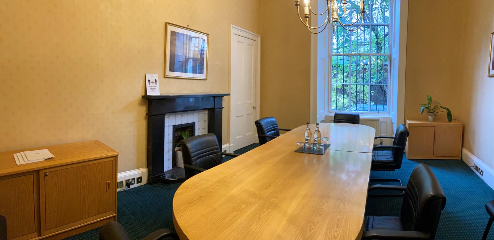 A conference room with a long wooden oval table surrounded by black chairs. A chandelier hangs above, with a fireplace and artwork on the beige walls. There are large windows with a view of greenery, and a cabinet on the left.
